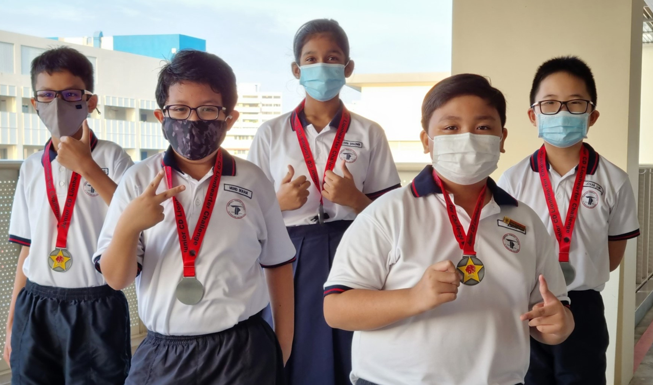 Five masked schoolchildren in uniforms proudly display star medals on red lanyards outdoors.